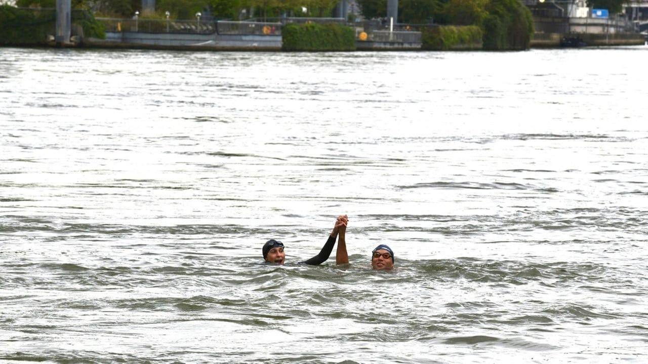 Eine Frau und ein Mann schwimmen in einem Fluss. Sie halten sich an den Händen und haben diese Hände hoch aus dem Wasser erhoben. 