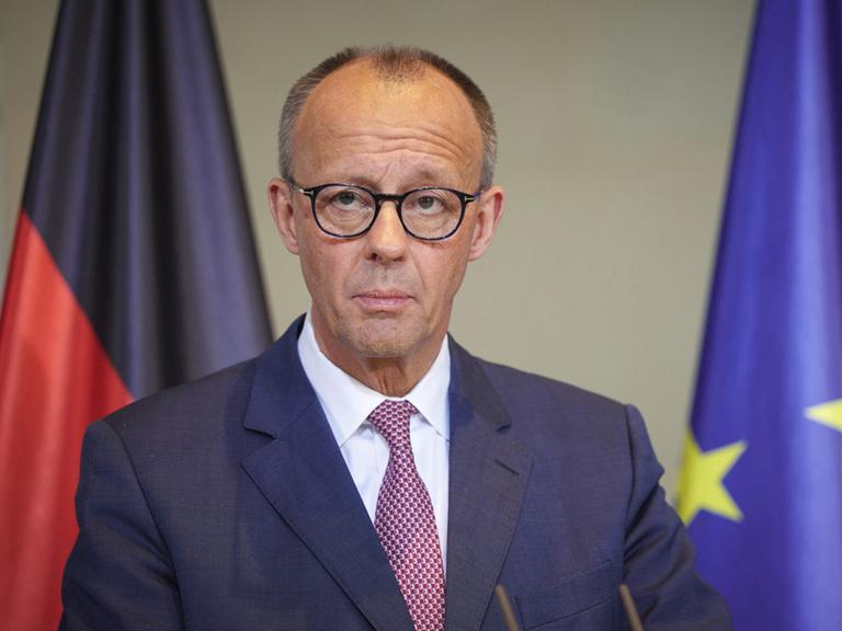 Bundeskanzler Friedrich Merz steht an einem Rednerpult, im Hintergrund sieht man im Anschnitt eine Deutschland- und eine Europa-Flagge.