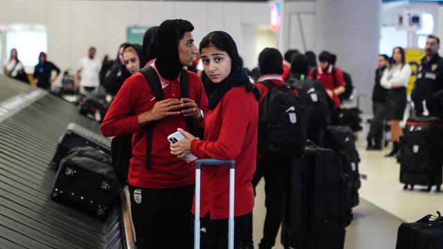 Zwei Frauen mit dunklen Kopftüchern und roten Pullovern stehen am Gepäck-Band von einem Flughafen. 