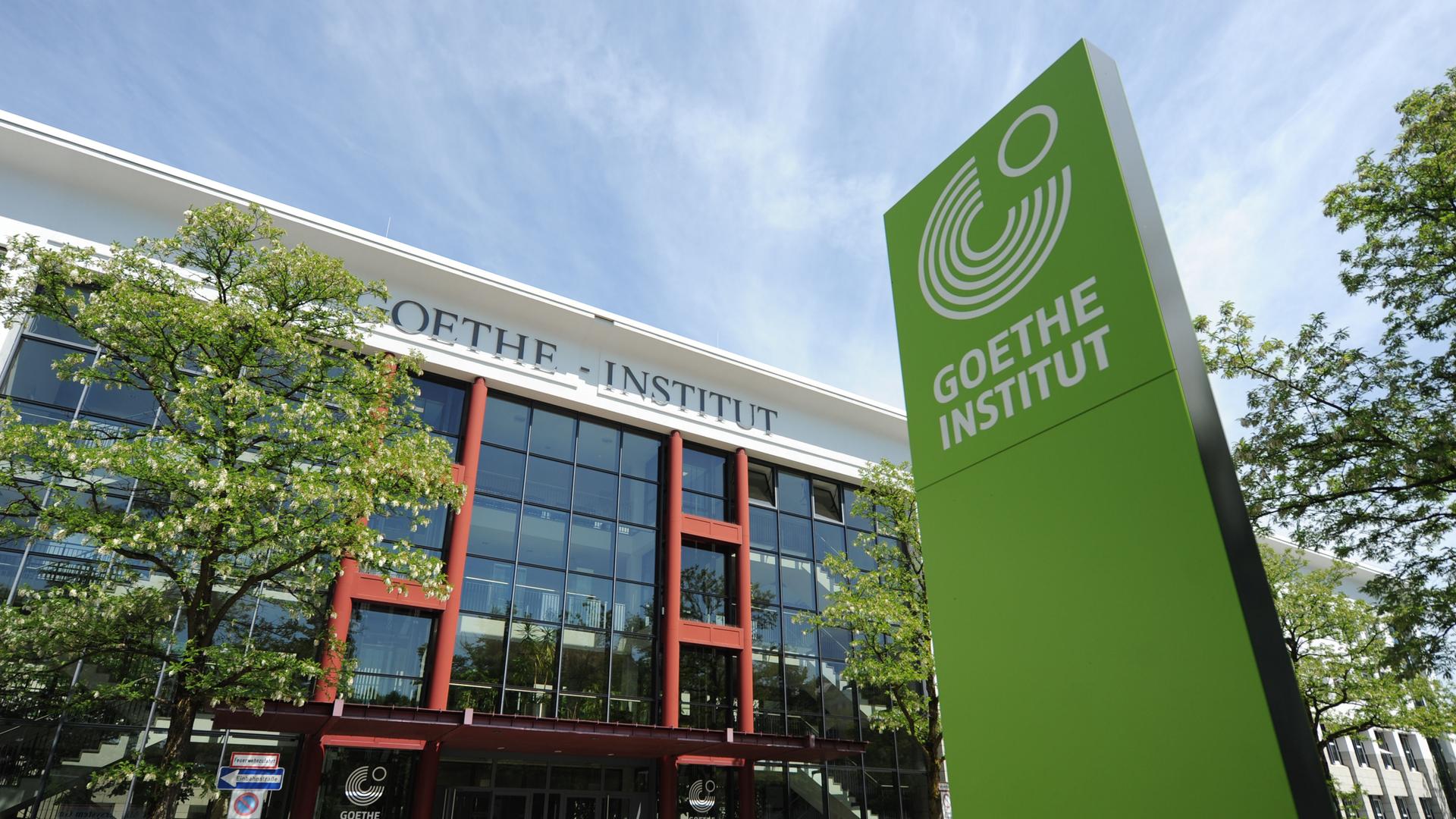 An der Zentrale des Goethe-Instituts in der Dachauer Straße 122 in München prangt oben ein Schriftzug "Goethe-Institut", vor dem Gebäude und im Bild rechts steht eine grünes Stele mit der Aufschrift "Goethe Institut" und dem Logo der Organisation, fünf Kreisen über 270 Grad, in der Aussparung oben rechts ist ein kleinerer Kreis kompett.