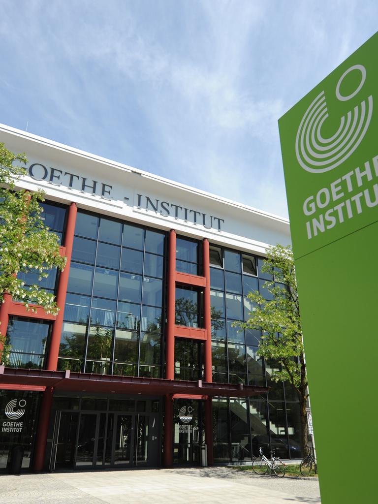 An der Zentrale des Goethe-Instituts in der Dachauer Straße 122 in München prangt oben ein Schriftzug "Goethe-Institut", vor dem Gebäude und im Bild rechts steht eine grünes Stele mit der Aufschrift "Goethe Institut" und dem Logo der Organisation, fünf Kreisen über 270 Grad, in der Aussparung oben rechts ist ein kleinerer Kreis kompett.