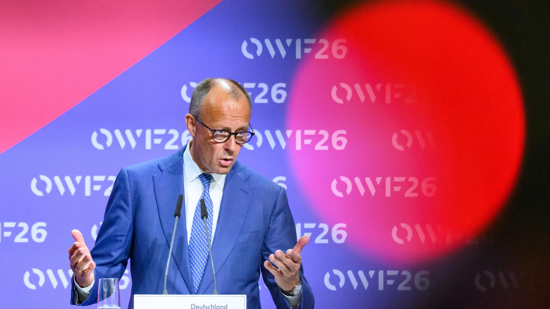 Friedrich Merz steht am Rednerpult vor rot-blauen Hintergrund mit OWF26-Logo. Er spricht gestikulierend. 