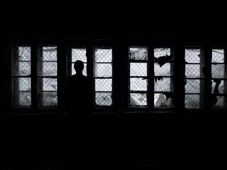 Eine Frau steht als Silhouette im Dunkeln vor vergitterten Fenstern und schaut hinaus