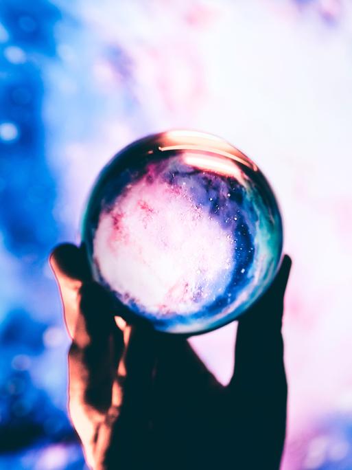 Nahaufnahme einer Hand, die vor blauem Hintergrund eine Glaskugel hält.