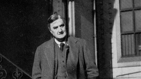 Eine Fotografie in schwarz weiß. Darauf zu sehen ist ein Mann, der einen langen Mantel trägt. Er steht vor einem Hauseingang mit Treppe. Es ist der englische Komponist Ralph Vaughan Williams.