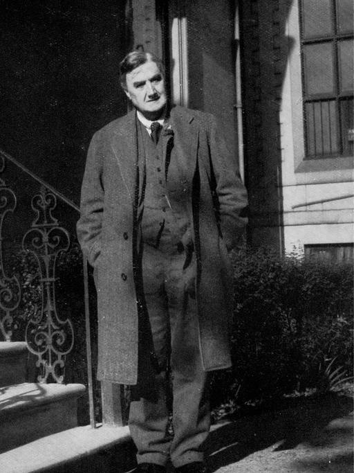 Eine Fotografie in schwarz weiß. Darauf zu sehen ist ein Mann, der einen langen Mantel trägt. Er steht vor einem Hauseingang mit Treppe. Es ist der englische Komponist Ralph Vaughan Williams.