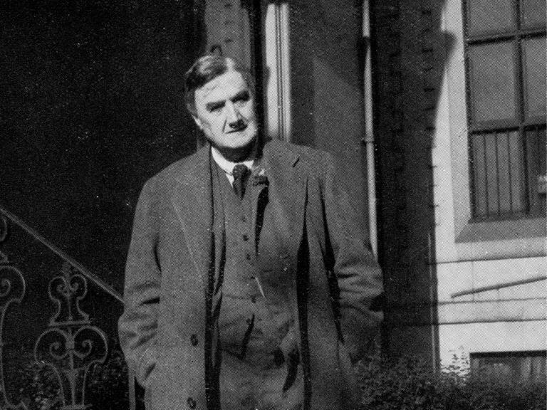 Eine Fotografie in schwarz weiß. Darauf zu sehen ist ein Mann, der einen langen Mantel trägt. Er steht vor einem Hauseingang mit Treppe. Es ist der englische Komponist Ralph Vaughan Williams.