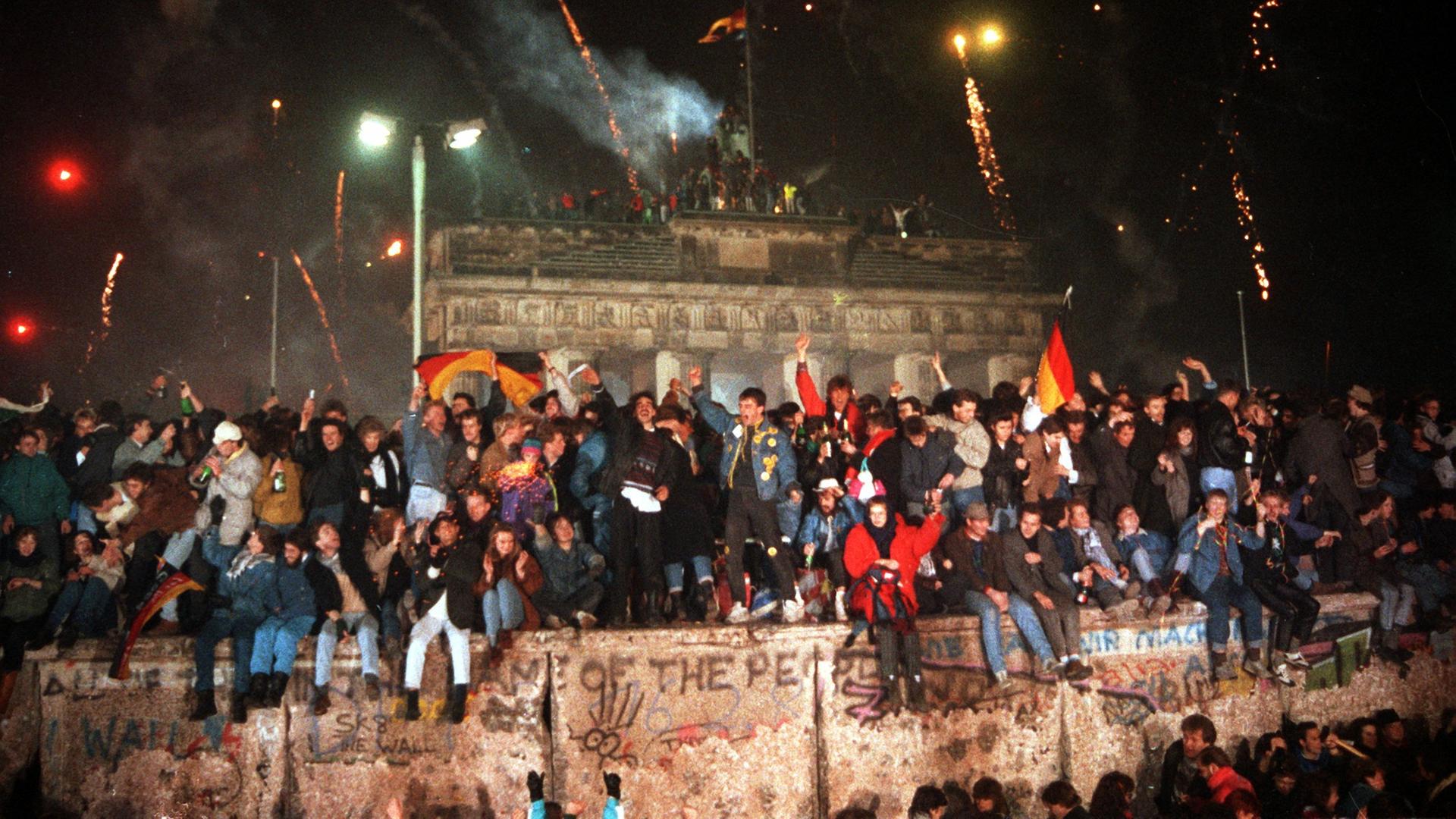 Jubelnd auf der Berliner Mauer vor dem Brandenburger Tor sitzend und stehend begrüßen Ost- wie Westberliner das Jahr 1990. Die erste deutsch-deutsche Silvesterfeier seit Öffnung der Mauer wurde mit einem großen Feuerwerk begangen.