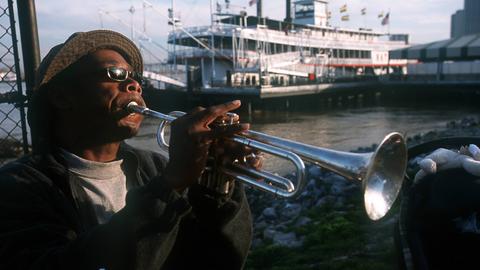Straßenmusiker in New Orleans, USA, spielt im Hafen Trompete. Im Hintergrund ist ein Ausflugsdampfer zu sehen.