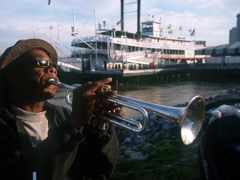 Straßenmusiker in New Orleans, USA, spielt im Hafen Trompete. Im Hintergrund ist ein Ausflugsdampfer zu sehen.