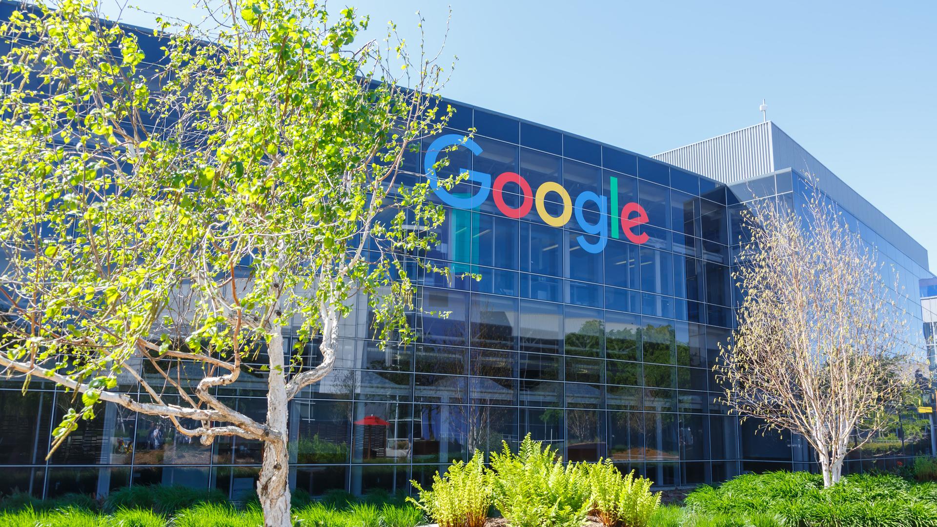 Die Google-Zentrale in Mountain View, Kalifornien. Ein Gebäude mit Glasfassade, davor Bäume.