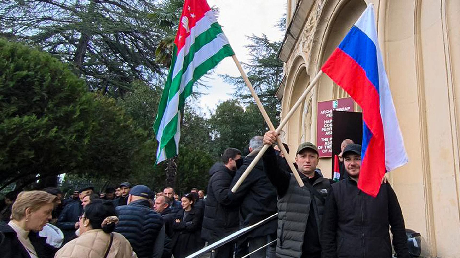 Die Demonstranten stehen vor dem Parlamentsgebäude. Im Vordergrund zwei Männer: Einer hält eine georgische Flagge und eine Abchasiens in einer Hand. 