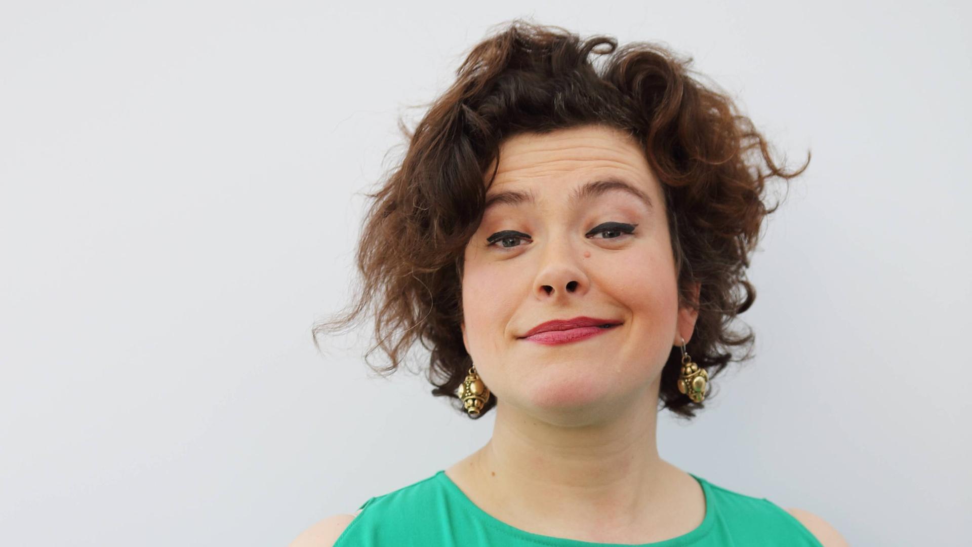 Nora Gomringer in einem türkis-grünen Kleid vor grauem Hintergrund. Sie hält ein Buch in der Hand und grinst in die Kamera. Nora Gomringer in einem türkis-grünen Kleid vor grauem Hintergrund. Sie hält ein Buch in der Hand und grinst in die Kamera.