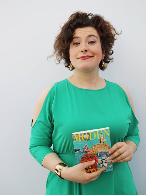 Nora Gomringer in einem türkis-grünen Kleid vor grauem Hintergrund. Sie hält ein Buch in der Hand und grinst in die Kamera.