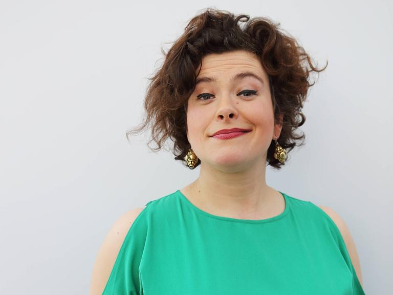 Nora Gomringer in einem türkis-grünen Kleid vor grauem Hintergrund. Sie hält ein Buch in der Hand und grinst in die Kamera.