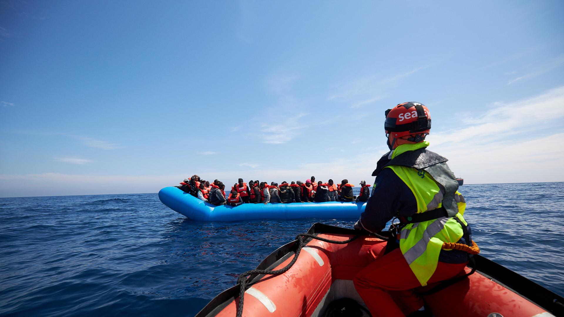 Seenotretter sitzt auf der Spitze eines Rettungsschlauchboots und steuert auf ein Schlauchboot mit Geflüchteten zu.