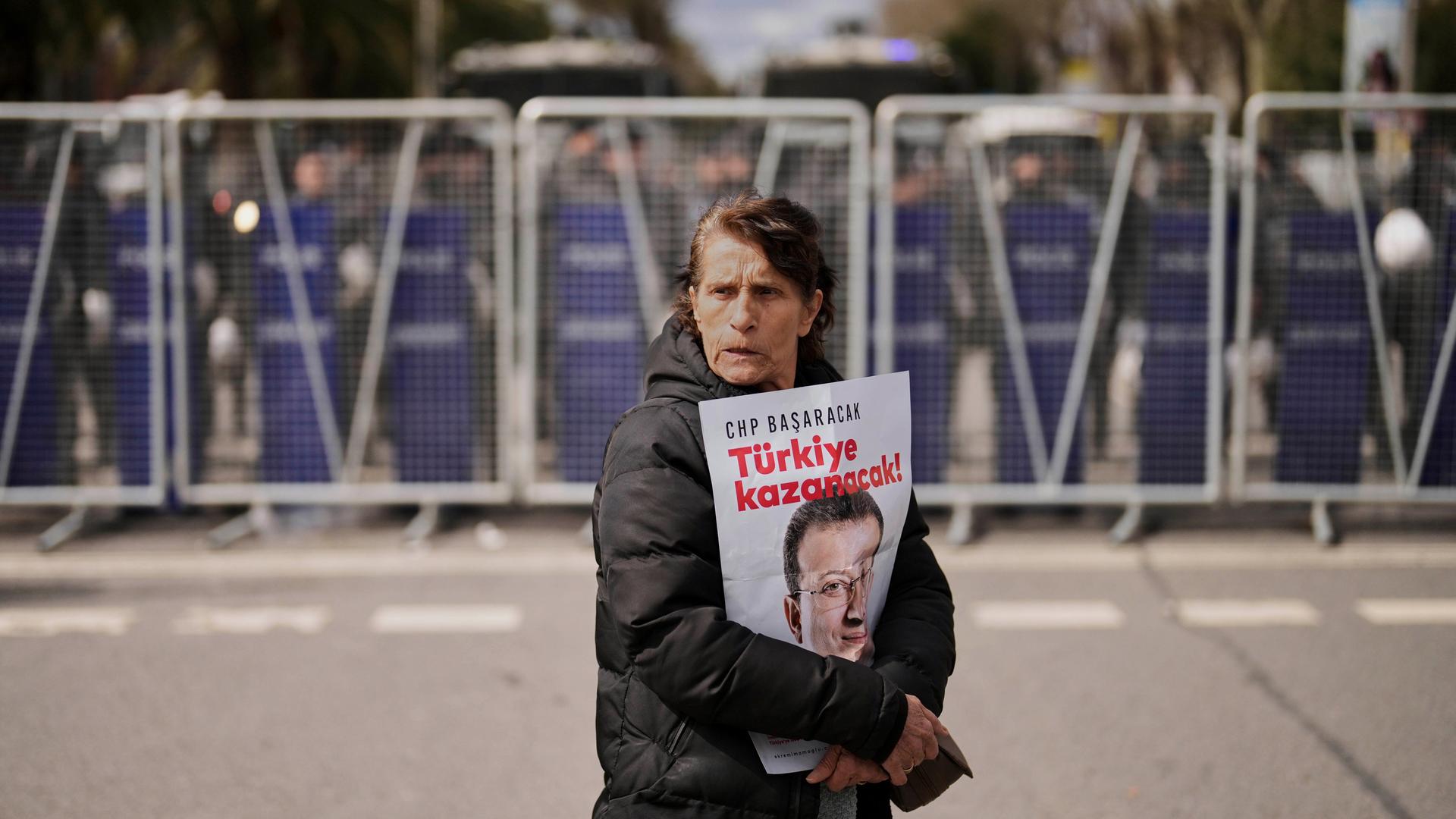 Eine Frau protestiert gegen die Festnahme des Istanbuler Bürgermeisters Imamoğlu. Sie hält ein Poster mit seinem Kopf fest umschlungen, im HIntergrund ist ein großes Aufgebot von Polizei mit Absperrgittern. 