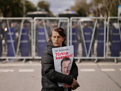 Eine Frau protestiert gegen die Festnahme des Istanbuler Bürgermeisters Imamoğlu. Sie hält ein Poster mit seinem Kopf fest umschlungen, im HIntergrund ist ein großes Aufgebot von Polizei mit Absperrgittern. 