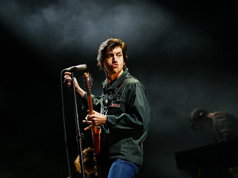 Arctic-Monkeys-Sänger Alex Turner posiert auf der Bühne mit Gitarre und Mikrofon.