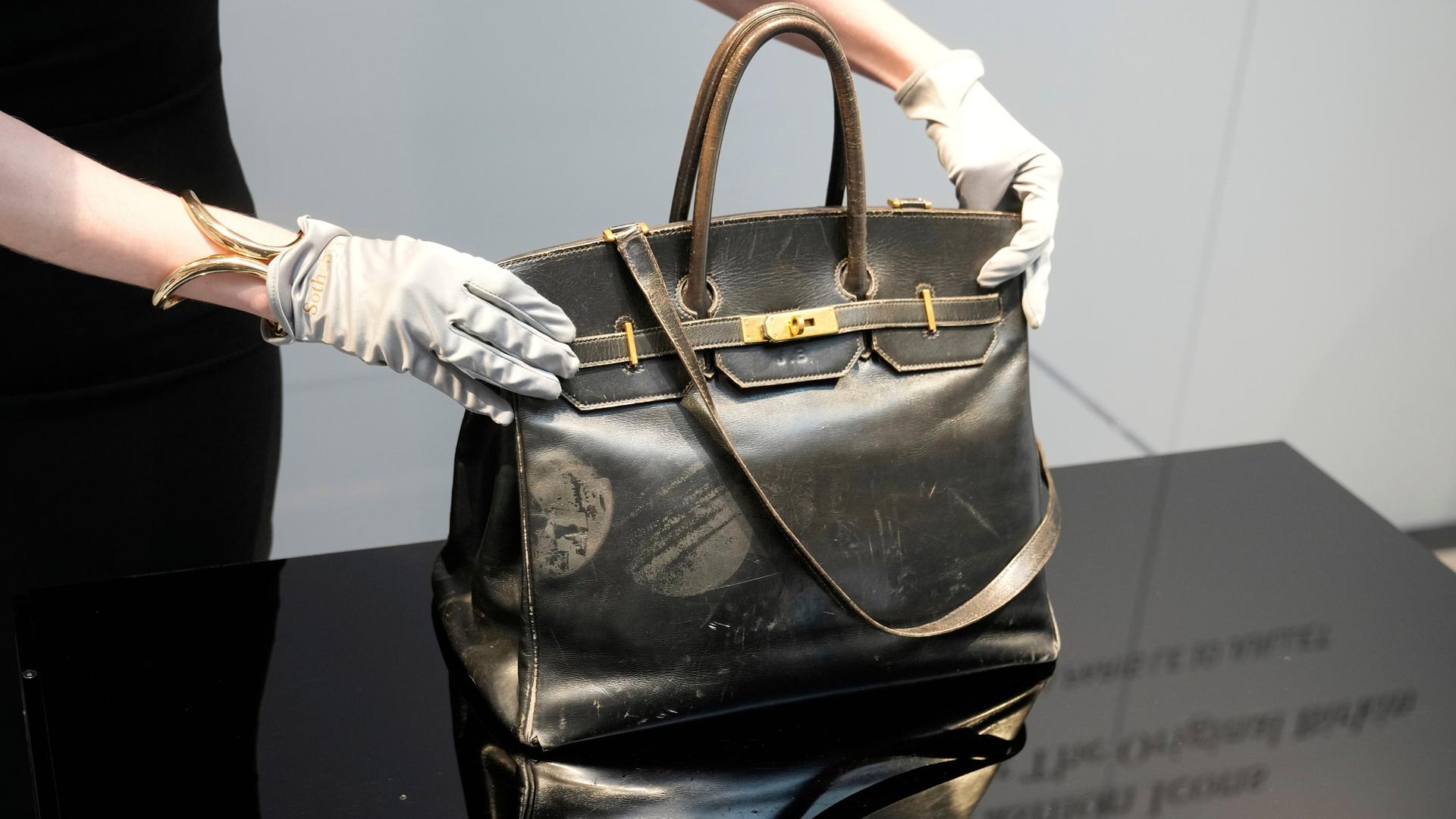 Zu sehen sind zwei Hände in weißen Handschuhen, die eine Designer-Handtasche auf einem Podest präsentieren. Zu sehen sind zwei Hände in weißen Handschuhen, die eine Designer-Handtasche auf einem Podest präsentieren.