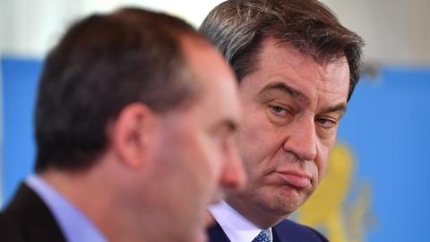 Das Bild zeigt die Köpfe der beiden: Aiwanger unscharf im Vordergrund und Söder scharf dahinter, beide stehen vor einer Wand mit dem bayerischen Wappen.