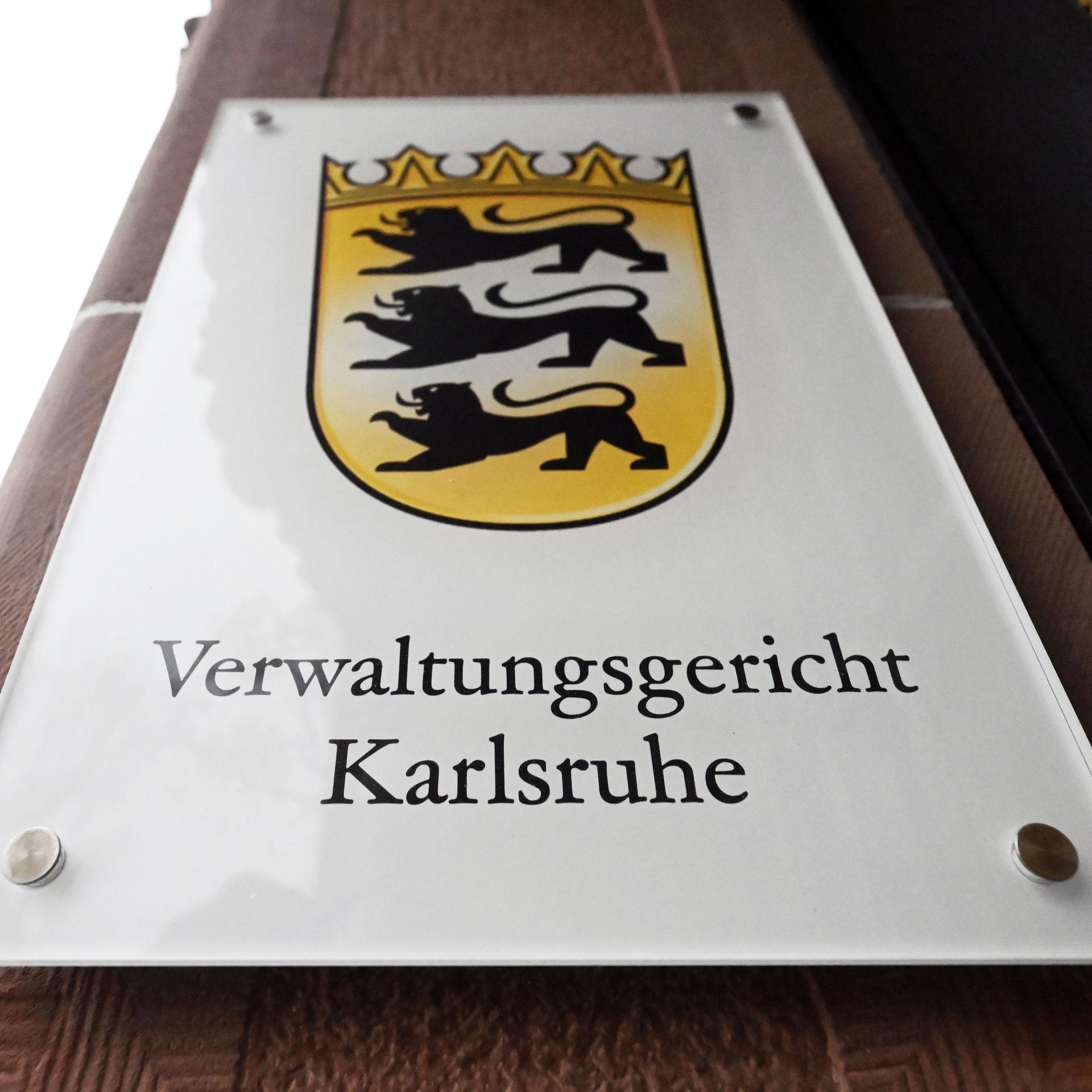 Ein Hinweisschild mit dem Landeswappen von Baden-Württemberg und dem Schriftzug „Verwaltungsgericht Karlsruhe“ am Eingang des Gerichtsgebäudes.
