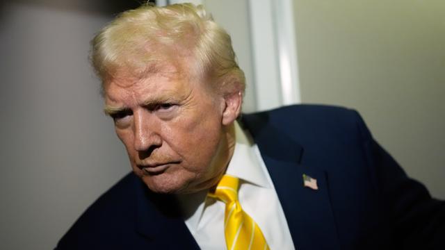 Donald Trump trägt einen dunklen Anzug und eine gelbe Krawatte. Er lehnt sich nach vorne und blickt kritisch.