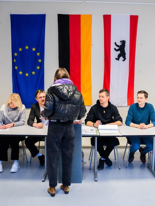 In einem Wahllokal sitzen Wahlhelfende an einem langen Tisch, vor dem eine Wählerin steht. An der Wand hängen die Europa-, die Deutschland- und die Berlinfahne.