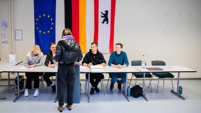 In einem Wahllokal sitzen Wahlhelfende an einem langen Tisch, vor dem eine Wählerin steht. An der Wand hängen die Europa-, die Deutschland- und die Berlinfahne.