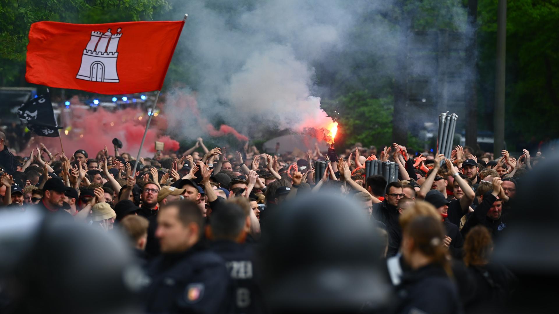 Aus einer dichten Menge von Fußball-Fans ragt eine St. Pauli-Fahne, es brennt Pyrotechnik. Dafür stehen Polizisten, zum Teil in Schutzkleidung.