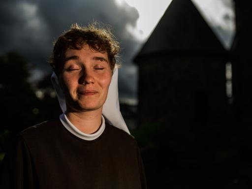Eine junge Frau mit Nonnenschleier sitzt lächelnd und mit geschlossenen Augen in einem Garten. Hinter ihr die Umrisse eines Klostergebäudes.