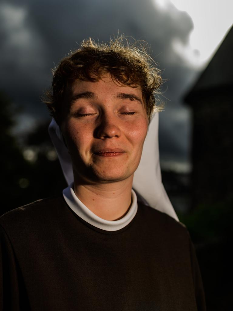 Eine junge Frau mit Nonnenschleier sitzt lächelnd und mit geschlossenen Augen in einem Garten. Hinter ihr die Umrisse eines Klostergebäudes.