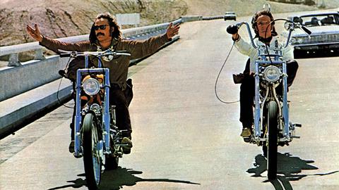 Dennis Hopper und Peter Fonda fahren in einer Filmszene mit Chopper-Motorrädern nebeneinander. Dennis Hopper (l.) fährt freihändig und breitet die Arme aus. 