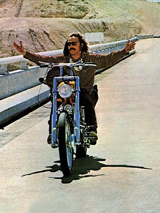 Dennis Hopper und Peter Fonda fahren in einer Filmszene mit Chopper-Motorrädern nebeneinander. Dennis Hopper (l.) fährt freihändig und breitet die Arme aus. 