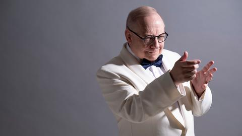 Ein Mann im weißen Anzug und runder, schwarzer Brille dirigiert in die Luft, mit vor der Brust erhobenen Händen. Er dreht sich zum rechten Bildrand und schaut konzentriert aus dem Bild hinaus. 
