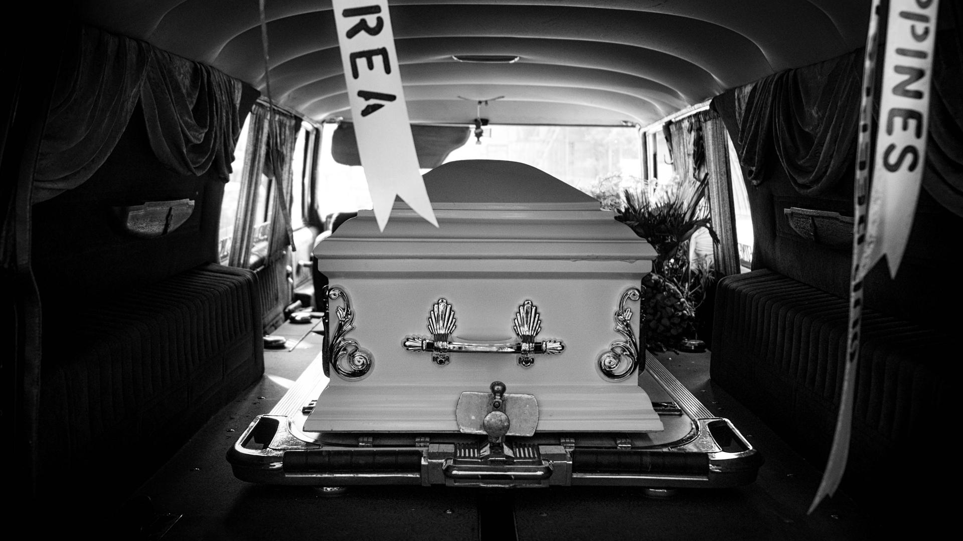 Schwarz-weiß-Bild: Blick in einen Leichenwagen mit offenem Kofferraum, darin ein weißer, edler Sarg. Schwarz-weiß-Bild: Blick in einen Leichenwagen mit offenem Kofferraum, darin ein weißer, edler Sarg.