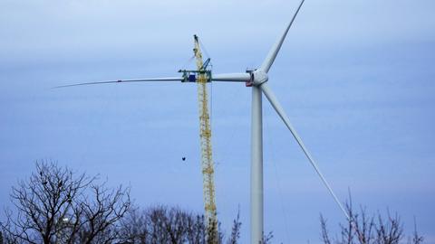 Ein Kran hebt ein Rotorblatt für eine neue Windenergieanlage des deutschen Herstellers Nordex nach oben. Zwei Rotorblätter sind bereits an der Anlage.