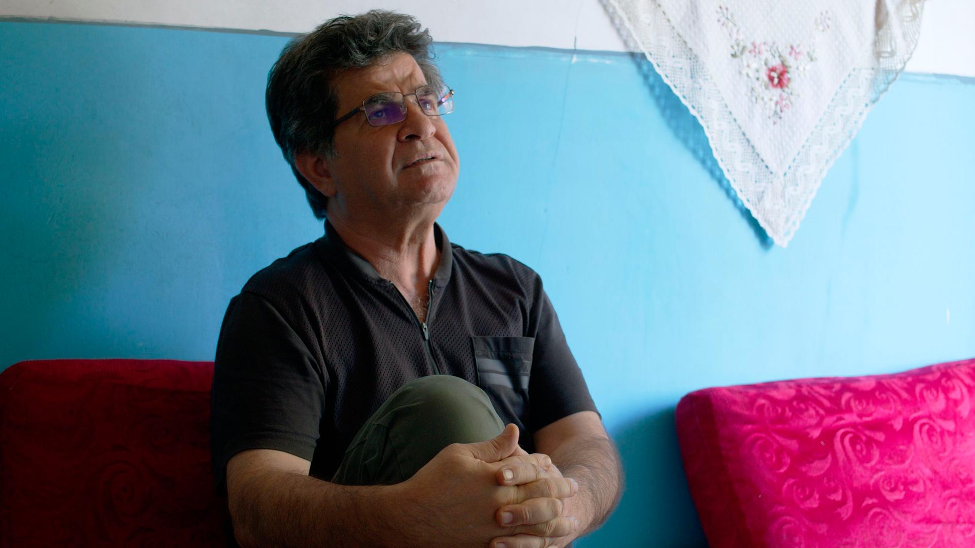 Der iranische Regisseur Jafar Panahi sitzt vor einer hellblauen Wand und hält sein rechtes Knie mit den Händen umschlungen. Der iranische Regisseur Jafar Panahi sitzt vor einer hellblauen Wand und hält sein rechtes Knie mit den Händen umschlungen.
