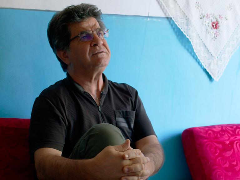 Der iranische Regisseur Jafar Panahi sitzt vor einer hellblauen Wand und hält sein rechtes Knie mit den Händen umschlungen.