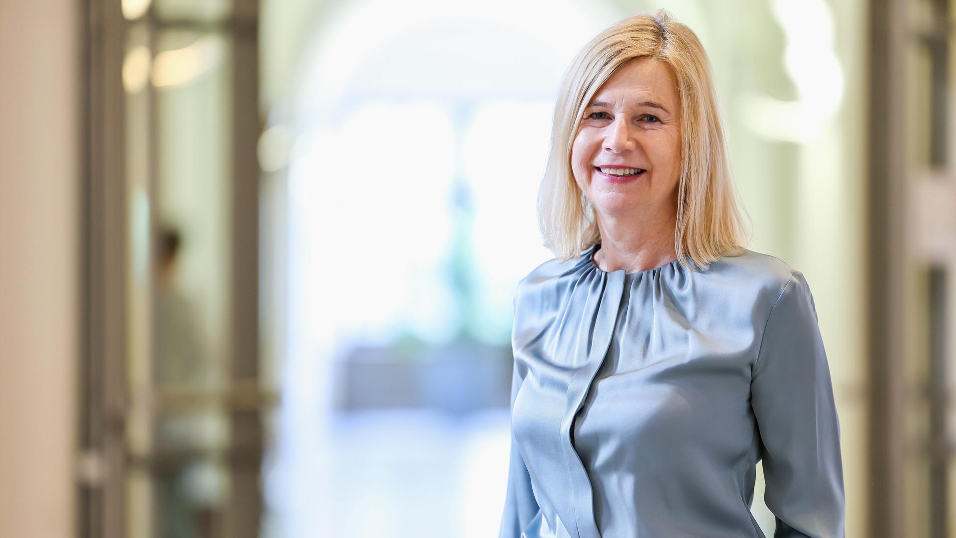 Angerer steht in einem Flur ihres bisherigen Gerichts. Sie trägt ihre blonden langen Haare offen über eine hellblaue Bluse und lächelt in die Kamera.