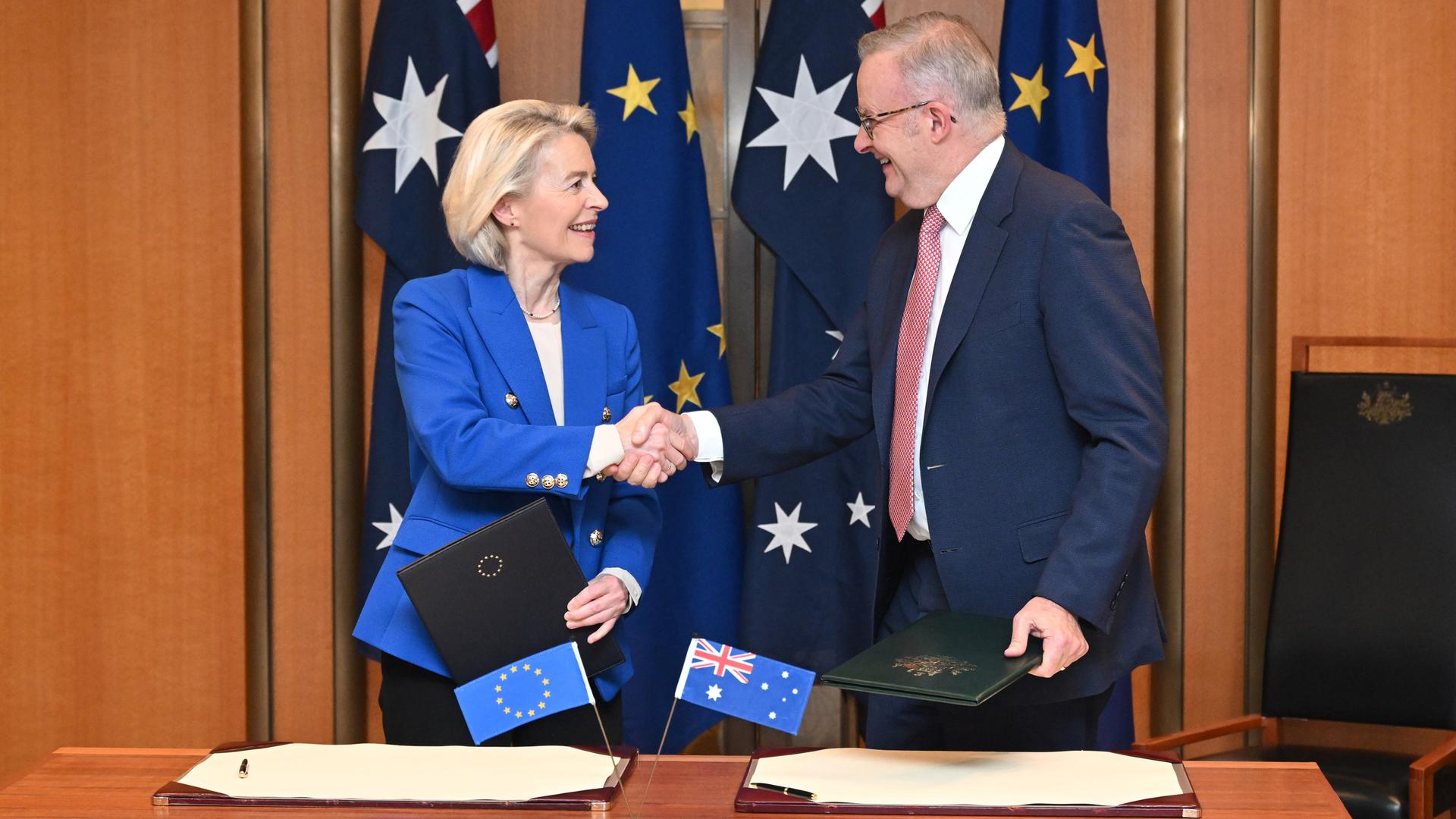 Die beiden stehen vor Fahnen beider Seiten und hinter einem Tisch. Sie schütteln sich die Hände und lächeln dabei.