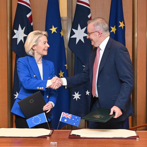 Die beiden stehen vor Fahnen beider Seiten und hinter einem Tisch. Sie schütteln sich die Hände und lächeln dabei.