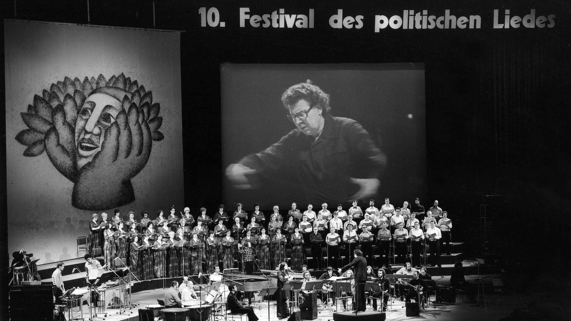 Schwarz-weiß-Foto der Bühne, auf dem eine Videoleinwand aufgebaut ist, auf der Dirigent für das große Auditorium sichtbar ist. 