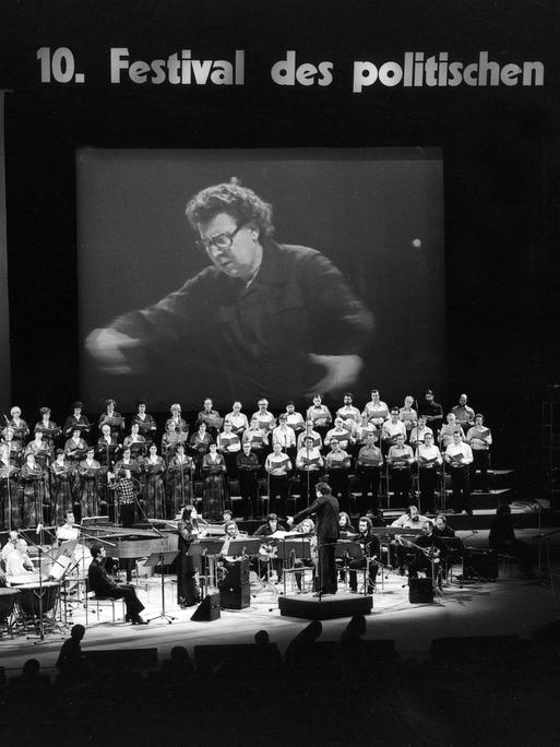 Schwarz-weiß-Foto der Bühne, auf dem eine Videoleinwand aufgebaut ist, auf der Dirigent für das große Auditorium sichtbar ist. 