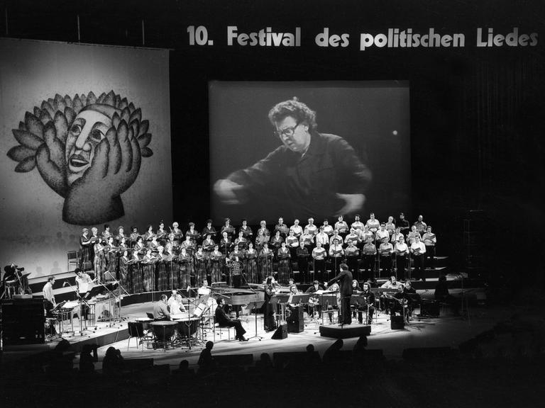 Schwarz-weiß-Foto der Bühne, auf dem eine Videoleinwand aufgebaut ist, auf der Dirigent für das große Auditorium sichtbar ist. 