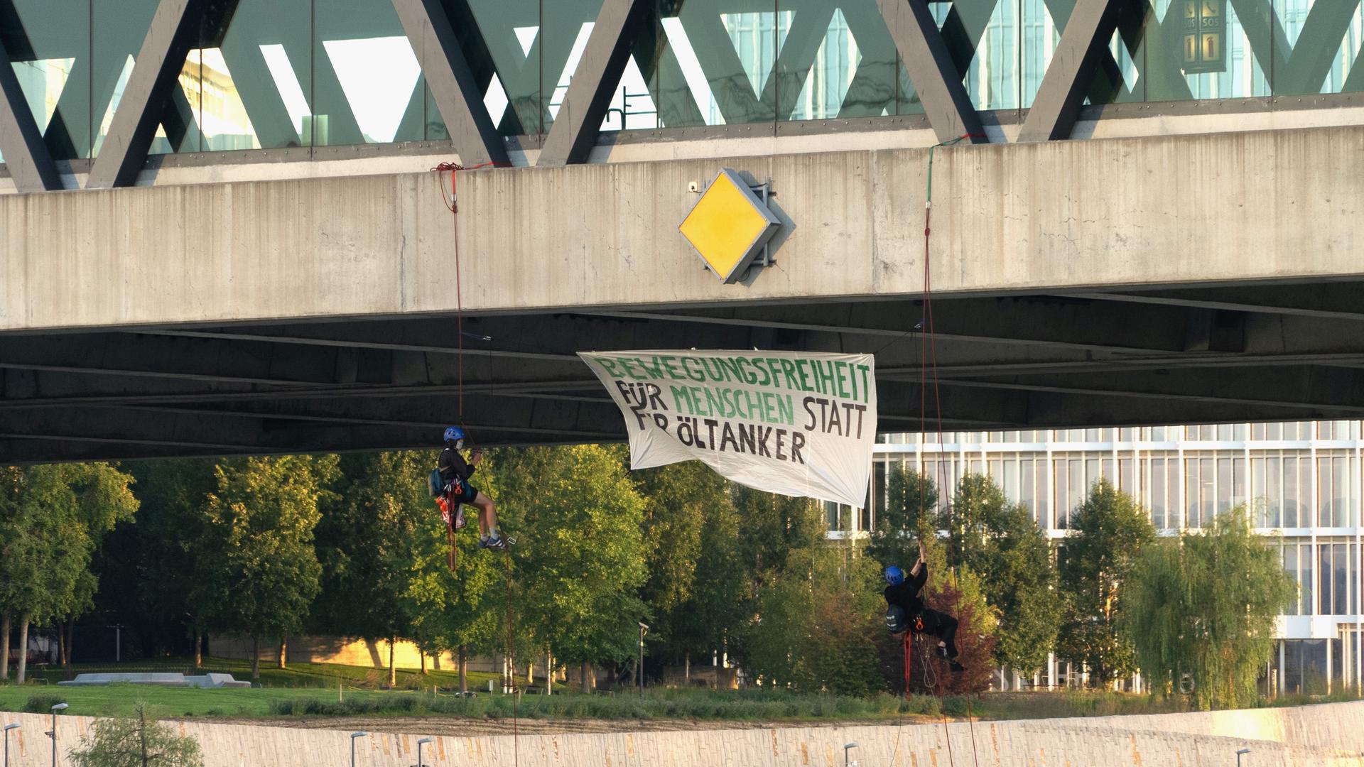 Klimaaktivistinnen und -aktivisten haben am frühen Freitagmorgen versucht den Schiffsverkehr bei der Dreirosenbrücke in Basel zu blockieren. Unter der Brücke hängt ein Mann an einem Seil und ein Plakat, auf dem steht "Bewegungsfreiheit für Menschen statt für Öltanker".
