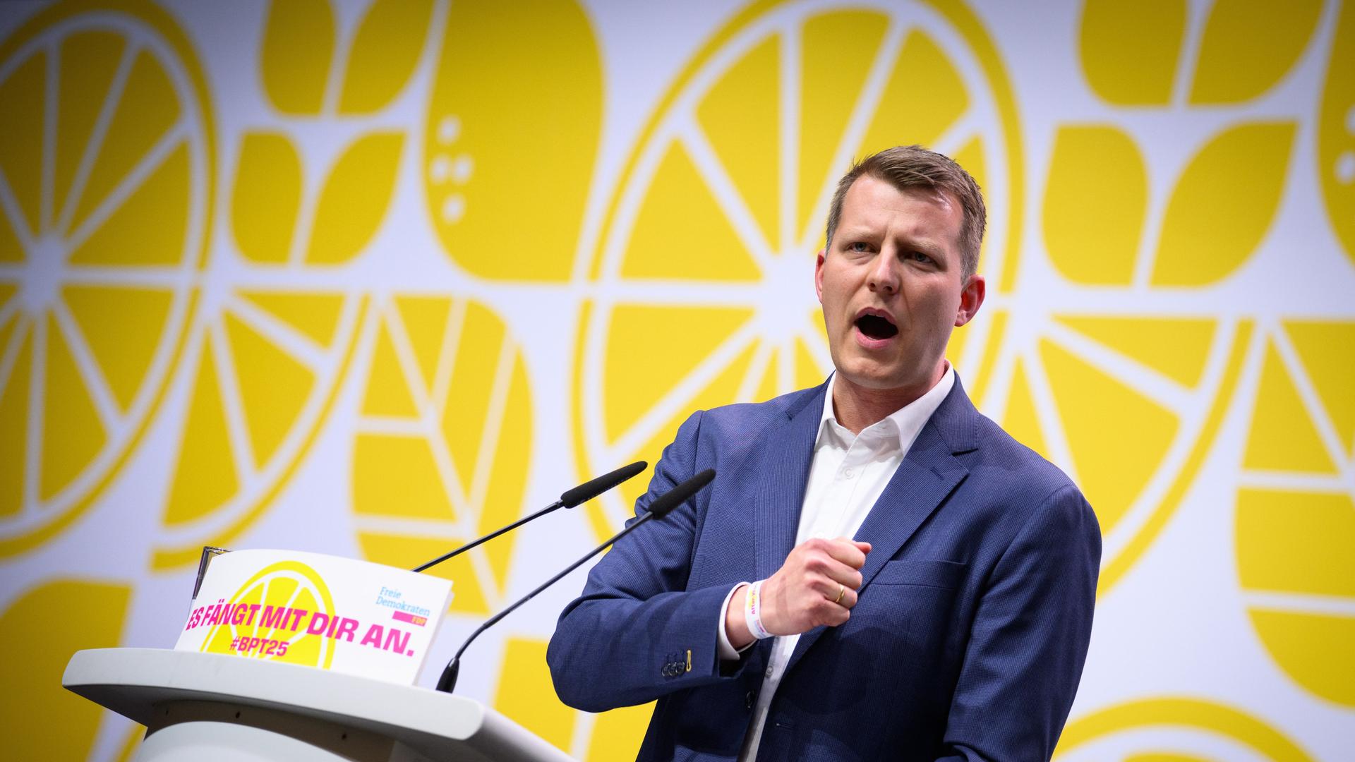 Henning Höne steht hinter einem Rednerpult der FDP. Er trägt einen blauen Anzug, darunter ein weißes Hemd und gestikuliert kämpferisch. Im Hintergrund ist eine Wand bedruckt mit gelben Zitronenscheiben zu sehen.