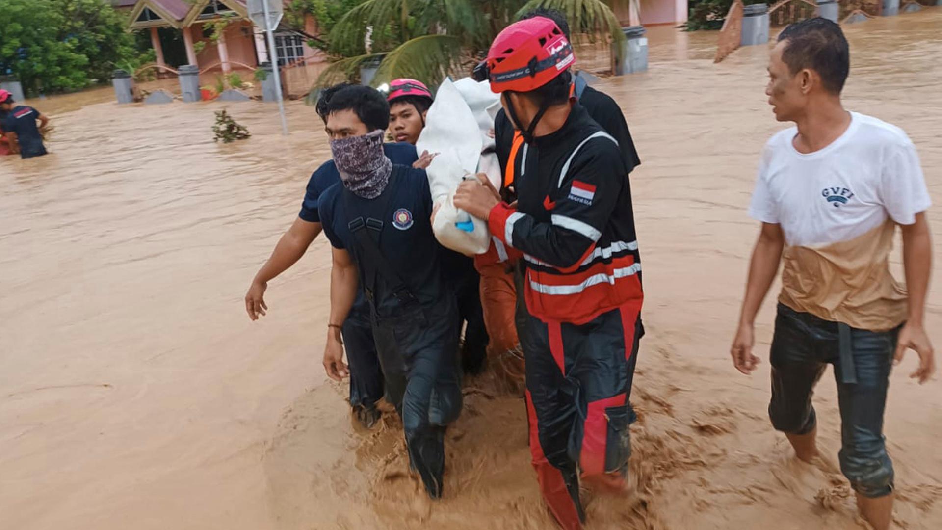 Rettungskräfte bringen Opfer einer Überschwemmung in Indonesien in Sicherheit. Alle Personen stehen bis zu den Knien im Wasser. Im Hintergrund sind Häuser.