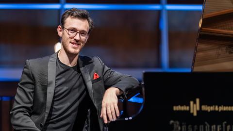 Aris Alexander Blettenberg sitzt in einem Anbzug mit rotem Einstecktuch am Flügel im Kammermusiksaal des Deutschlandfunks in Köln.