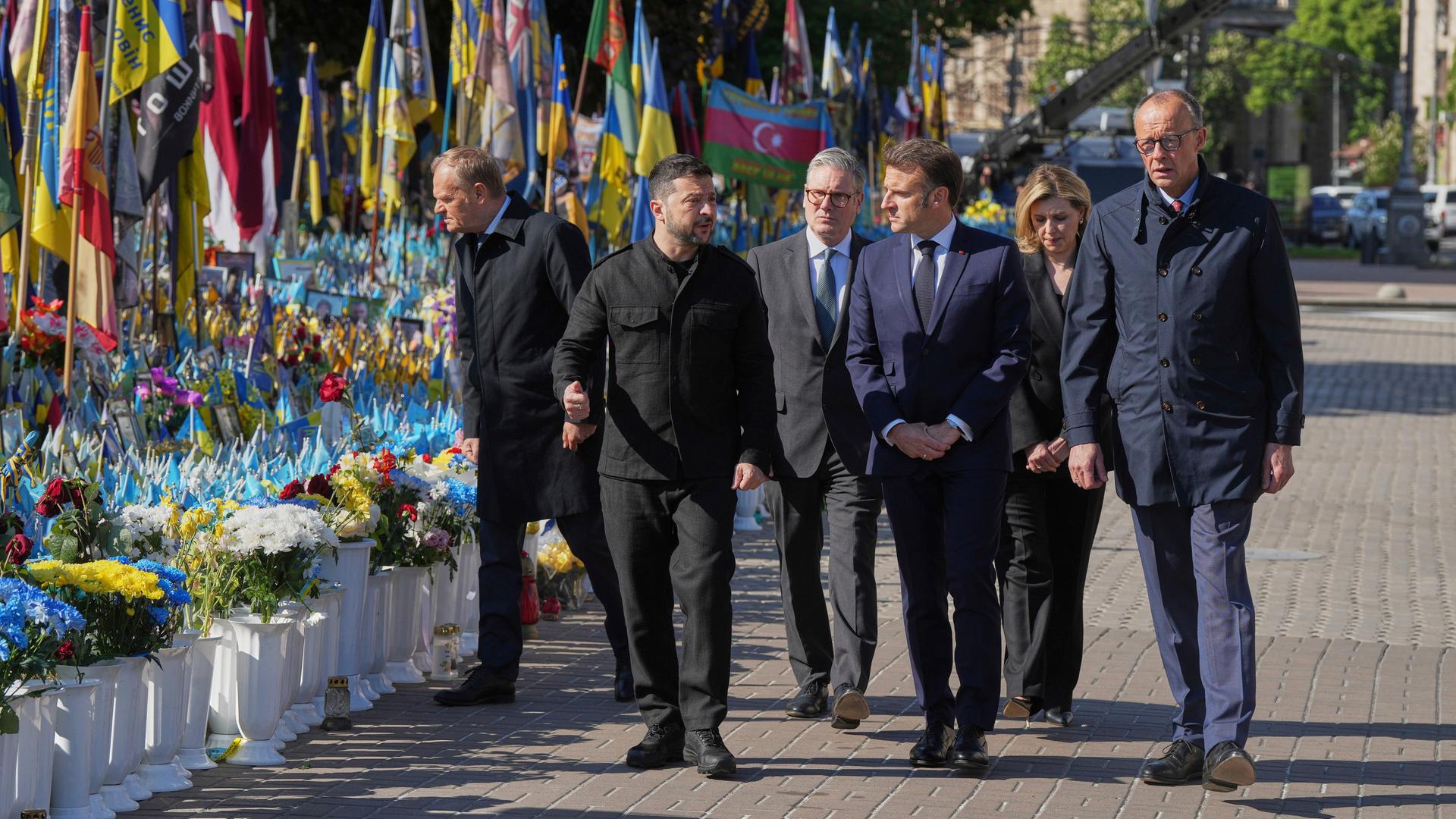 Donald Tusk, Wolodymyr Selenskyj, Keir Starmer, Emmanuel Macron und Friedrich Merz gehen an einem mit Flaggen und Blumen geschmückten Ort vorbei.
